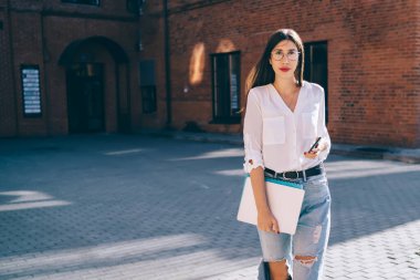 Modern cep telefonuyla üniversite kampüsünde yürüyen ve aynı anda kameraya bakan güzel bayan öğrencinin tam boy portresi. Moda kıyafetleri içinde çekici bir hippi kız.