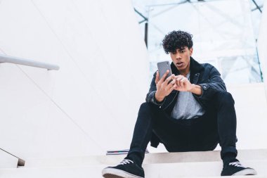 Kafası karışmış, şık giyinmiş, kıvırcık saçlı, beyaz, modern bir binada merdivenlerde otururken akıllı telefon kullanan Afro-Amerikan bir adam.