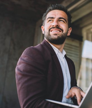 Sakallı, takım elbiseli, dizüstü bilgisayarı karıştıran ve modern binanın önünde gülümseyerek duran İspanyol bir adam.
