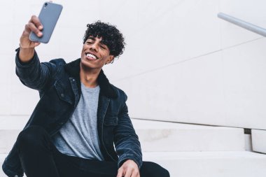 Şık takım elbiseli mutlu kıvırcık siyah adam elinde akıllı telefonuyla büyük beyaz odada oturmuş selfie çekiyordu.