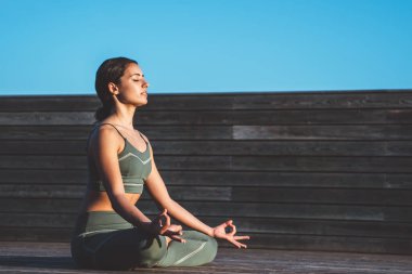 Yeşil spor elbiseli, sakin etnik zayıf bir kadının Lotus duruşu içinde el ele tutuşup ahşap terasta gözlerini kapamasının yan görüntüsü.