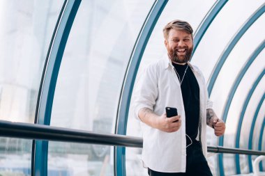 Kolları kulaklıklı, beyaz tişörtlü mutlu sakallı adam yaya köprüsünde akıllı telefondan müzik dinliyor.