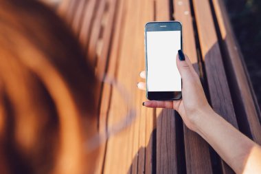 Kırpılmış, kızıl saçlı, sıradan giyinmiş, telefon kullanarak sosyal medyada arkadaşlarıyla sohbet eden, ahşap bankta oturan kadınların arka plan görüntüsü.