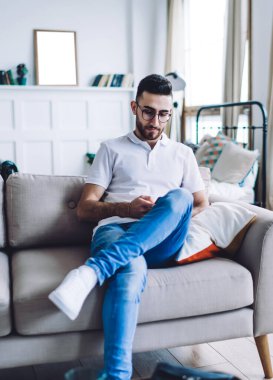 Kot pantolon ve beyaz polo tişörtlü genç adam gri rahat koltukta oturuyor, bacak bacak üstüne atıyor ve arka planda bulanık bir oturma odasıyla akıllı telefondan mesajlaşıyor.