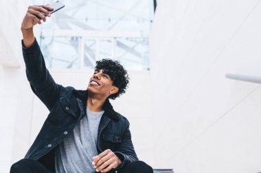 Şık kot elbiseli mutlu kıvırcık Afrikalı Amerikalı. Akıllı telefonlu, büyük beyaz bir odada merdivenlerde oturmuş, selfie çekiyor ve gülümsüyor.