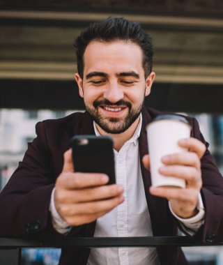 Neşeli Kafkas erkek milenyumu şık ceketli cep telefonuyla sohbet eden zengin iş adamı dışarıda kahve molası veren akıllı telefonuyla sosyal ağları okuyarak eğleniyor.