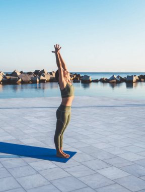 Yeşil spor kıyafetli zayıf bir kadın yoga yapıyor ve sabah suyun yakınındaki toprak setinde mavi hasırın üzerinde dağ pozu veriyor.