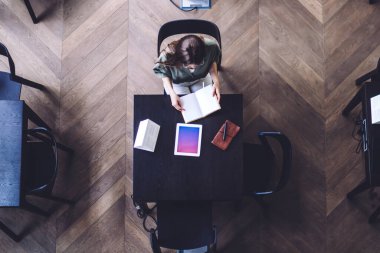 Yukarıdan gelen kadın, elinde kitap ve defterle dijital tabletin yanında oturmuş, gradyan ekranlı ve üniversite kütüphanesinde kitap okuyor.