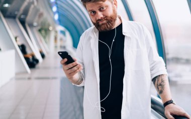 Elektronik kulaklıklı yakışıklı hippi adamın yarım boy portresi. Kapalı mekanda kameraya bakıyor ve öğrenmek için ses kitabı dinliyor. Cep telefonlu, boş zamanlarında poz veren sakallı bir adam.