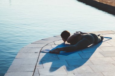 Yoga kıyafetleri içinde tanınmayan bir kadın. Bacaklarını geniş bacaklarıyla öne doğru uzatıyor. Elleri arkada, taş iskelede.