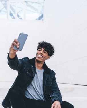 Şık takım elbiseli mutlu kıvırcık siyah adam elinde akıllı telefonuyla büyük beyaz odada oturmuş selfie çekiyordu.