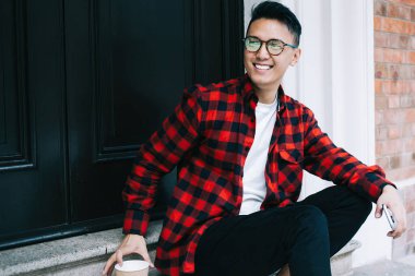 Tatmin olmuş Asyalı ekose gömlekli ve gözlüklü genç bir adam. Kapıda vakit geçirip kahvenin tadını çıkarıyor ve gülümsüyor ve başka tarafa bakıyor.