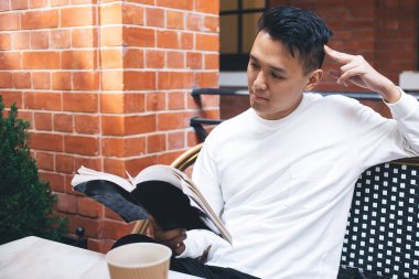 Beyaz gömlekli düşünceli, modern Asyalı bir erkek bir fincan kahveyle kafede oturuyor ve favori yazarının heyecan verici dedektif hikâyesini okuyor.