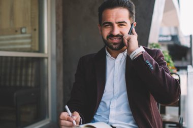 Resmi olarak İspanyol sakallı bir adam modern binanın balkonunda otururken ve akıllı telefondan konuşurken planlamacı gibi yazılar yazar ve kameraya bakar.