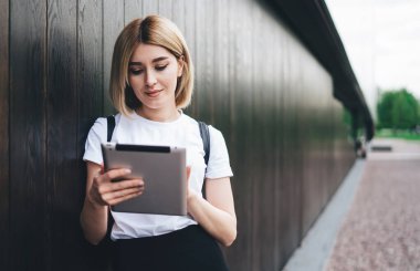 Sıradan kıyafetler giymiş genç sarışın bir kız öğrenci. Tableti karıştırıyor ve dinleniyor. Sokaklarda durup ahşap duvara yaslanıyor.