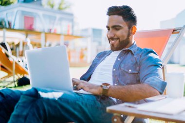 Gündelik kıyafetlerle gezen mutlu etnik adam dizüstü bilgisayarla yazı yazarak parktaki sandalyede oturarak yeni bir başlangıç yapıyor.