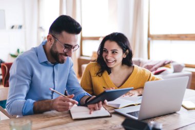 Gülen çift, günlük giysiler giyip not defterine notlar yazarken tablet ve dizüstü bilgisayarları karıştırıyor ve evdeki masada oturuyor.