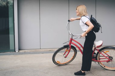 Modern binanın dış tarafına bakan pembe bisikletle gezerken kısa saçlı, şık sarışın bir kadının yan görünüşü.