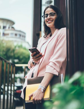Kağıt klasörü ve günlüğü olan, cep telefonu uygulaması kullanan ve yaz günü binanın kapısında dururken gülümseyen modern başarılı bir kadından.