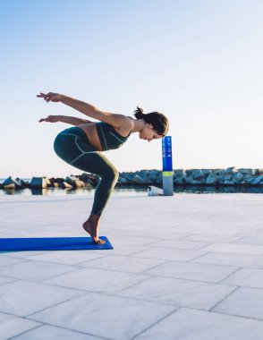 Yeşil spor kıyafetli zayıf bir kadın parmak uçlarında mavi minder üzerinde duruyor ve sabahları sette yoga yaparken dengede duruyor.