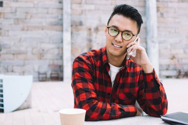 Düşünceli, genç, düşünceli, gözlüklü ve kareli tişörtlü Asyalı adam dışarıda, şehir masasında telefonla konuşurken başka tarafa bakıyor.