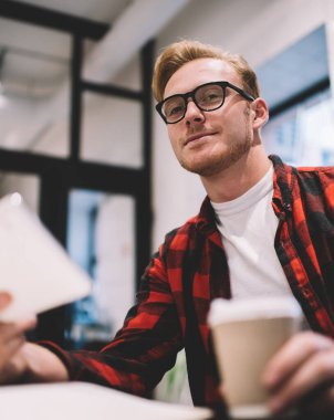Gündelik giysiler ve gözlüklerle yakışıklı genç adam kahve dükkanında kameraya bakarak bir kupa taze içecek ve telefon alıyor.