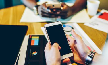 Yukarıdan gelen çok ırklı gözlüklü meslektaşlar cep telefonlarıyla sörf yapıyor ve masada dizüstü bilgisayar ve tabletle çalışma sürecini analiz ediyorlar.