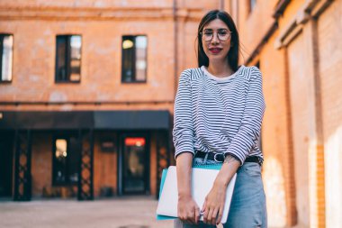 Serbest çalışmak için modern dizüstü bilgisayarı tutan ve boş zamanlarında açık havada kameraya bakan gündelik kıyafetli başarılı bir kadının yarım boy portresi.