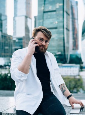 Gündelik giysiler ve yuvarlak gözlükler içinde odaklanmış bir erkek akıllı telefondan iş arkadaşıyla konuşuyor. Proje üzerinde çalışıyor. Modern şehrin sokaklarında dizüstü bilgisayarla kameraya bakıyor.