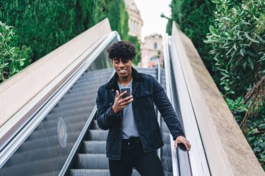 Moda kıyafetli pozitif hipster adam şehir yürüyen merdiveninde aşağı iniyor ve takipçilerle çevrimiçi iletişim kurmak için modern cep telefonu aygıtı kullanıyor, medya dosyalarını paylaşmak için neşeli erkek blogger ağı