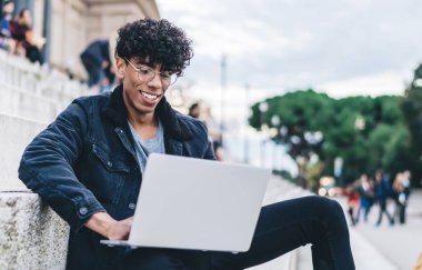 Gözlüklü mutlu etnik adam sıradan kıyafetler giyiyor. Şehir sokaklarında merdivenlerde arka planda insanlarla otururken bilgisayarını faiziyle kullanıyor.