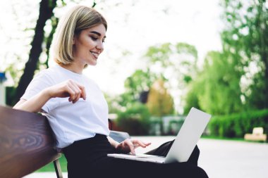 Neşe dolu ve iyimser bir kadın, parkta bankta oturmuş kahve içerken ve sosyal medyada gezinirken gülümsüyor.