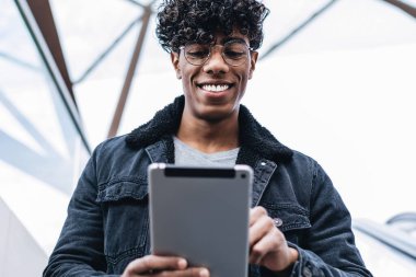 Optik gözlüklü neşeli Latin adam yürüyen merdivende aşağı iniyor ve dokunmatik ped aracılığıyla online sohbetten heyecanlanıyor, milenyum pozitif erkek blogcu mesajlar alıyor