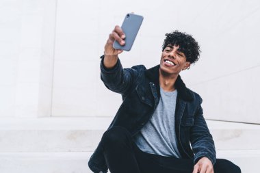 Şık takım elbiseli mutlu kıvırcık siyah adam elinde akıllı telefonuyla büyük beyaz odada oturmuş selfie çekiyordu.