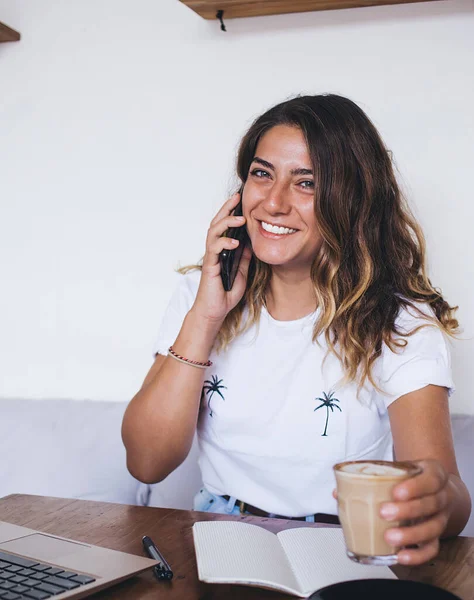 魅力的な幸せな笑顔の若い女性とコーヒーを片手にノートパソコンの前のカフェに座って電話で話す — ストック写真