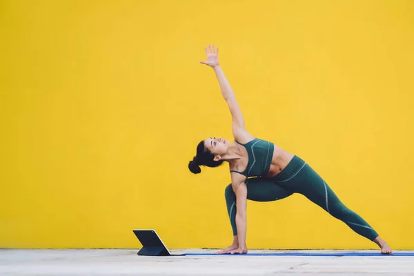 Adult slim female in tracksuit and with black hair standing on mat practicing yoga in parsvakonasana pose learning from online class while using tablet in yard on background of empty yellow wall