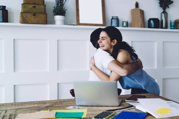 Side view of man in white T-shirt and young happy dark haired woman with toothy smile hugging and congratulating each other in modern cozy workplace