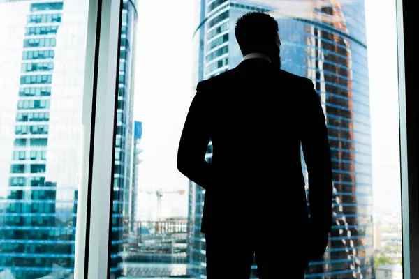 From below back view of faceless confident man in suit standing in darkness against office window looking away on skyscrapers