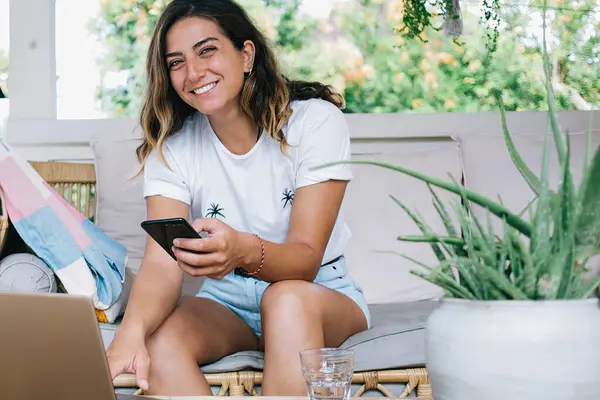 黒い髪の若い陽気な女性は スマートフォンを入力し ラップトップを使用している間 水のガラスとテーブルの近くの快適なソファに座って歯の笑顔で — ストック写真