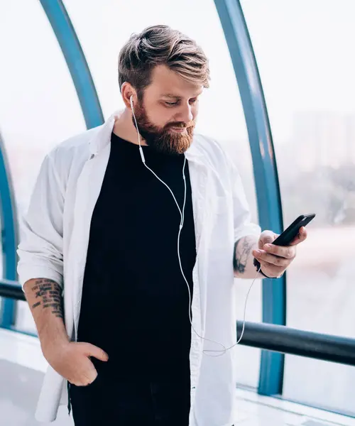 Man 30s checking received email from colleague while standing indoors and listening new music using playlist via electronic earphones, hipster guy received reading sms and choosing player song