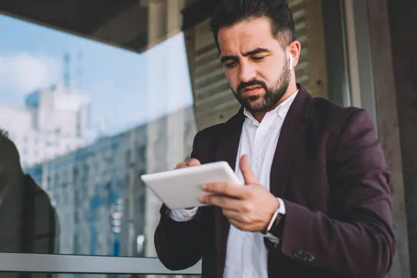 Pensive businessman in formal wear concentrated on making money transaction using digital tablet application outdoors, serious male entrepreneur checking news from exchange browse web page on touchpad