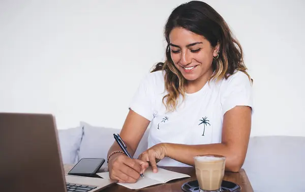 ノートパソコンとコーヒーを片手にカフェで小さなテーブルに座っている間 仕事のスケジュールやノートブックで書くことを計画している幸せな創造的な女性 — ストック写真