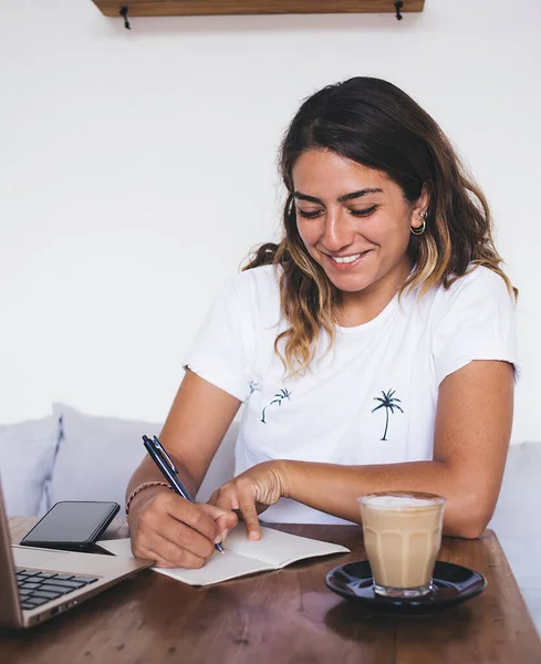 ノートパソコンとコーヒーを片手にカフェで小さなテーブルに座っている間 仕事のスケジュールやノートブックで書くことを計画している幸せな創造的な女性 — ストック写真