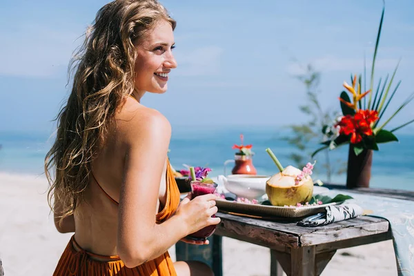 Young female tourist in summer orange gown drinking fruit smoothie after breakfast enjoying in views and looking away with blue sky on background