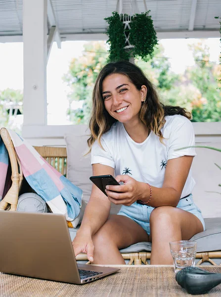 黒い髪の若い陽気な女性は スマートフォンを入力し ラップトップを使用している間 水のガラスとテーブルの近くの快適なソファに座って歯の笑顔で — ストック写真
