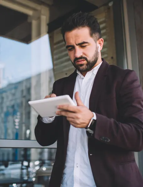 Pensive businessman in formal wear concentrated on making money transaction using digital tablet application outdoors, serious male entrepreneur checking news from exchange browse web page on touchpad