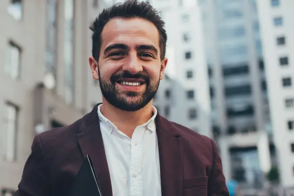 Half length portrait of cheerful caucasian guy in trendy outfit standing on urban setting background,young businessman holding paper folder and looking at camera satisfied with successful career
