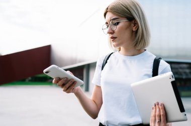 Gözlüklü, günlük giysiler içinde dikkatli bir kadın. Ofis binasının yanındaki caddede dikilirken konsantrasyonuyla akıllı telefona göz atıyor.