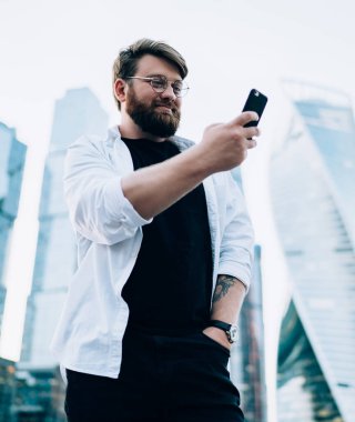 Düşük açılı mutlu, dolgun gözlüklü, sıradan kıyafetler giymiş, selfie yapan, arka planda modern ayna gökdelenlerinin karşısında duran bir adam.