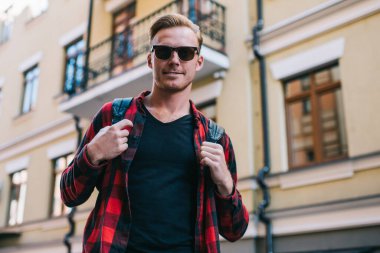 Güneş gözlüklü ve ekose gömlekli karizmatik neşeli bir adam. Sırt çantasıyla kameraya bakıyor ve şehirdeki çok katlı binaların bulanık dış görünüşlerine gülümsüyor.